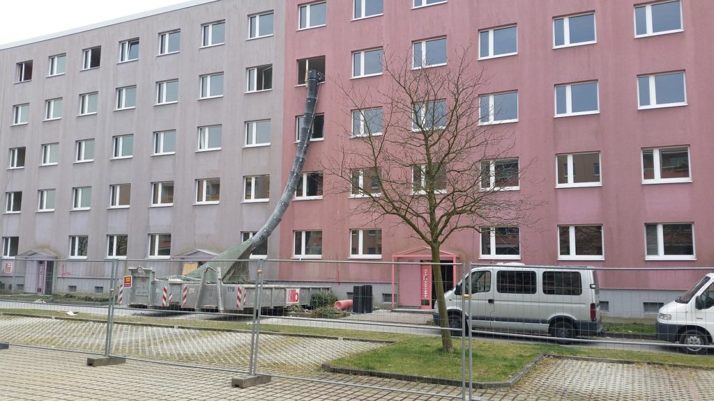 Außenansicht des Wohnblocks in der Irkutsker Straße vor der Sanierung. Aus einem Fenster führt eine Schuttrutsche in einen großen Container.