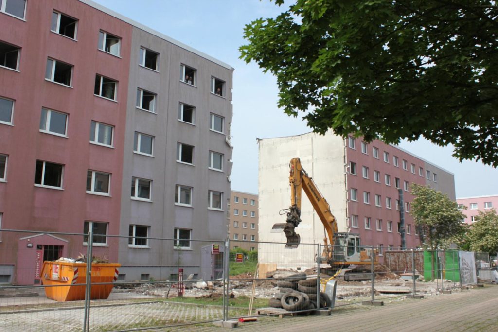 Der Mittelteil des Blocks wurde abgerissen. Davor steht noch ein Bagger und Arbeiter sehen aus einem Fenster des Nebenaufganges.